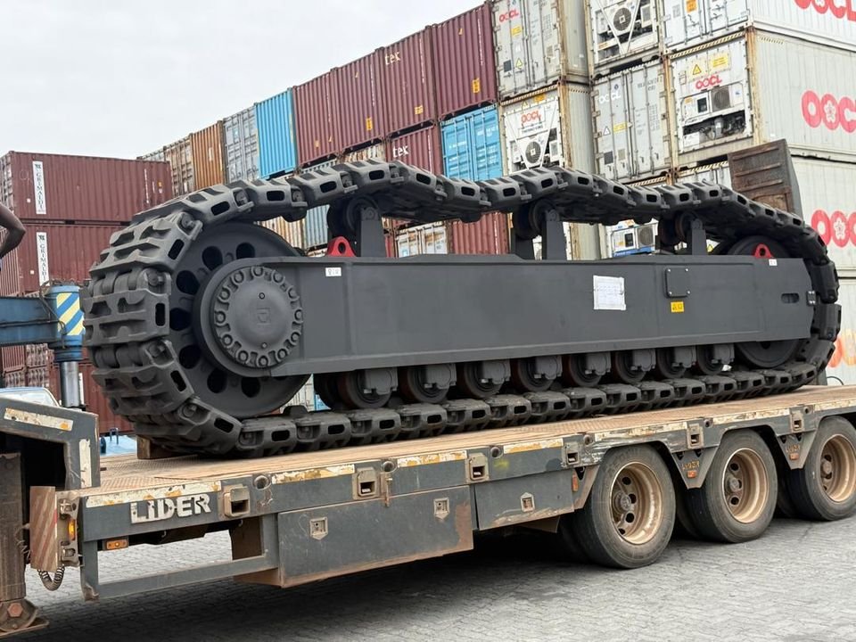 Crawler track frame loaded on a Lider lowbed trailer in front of stacked containers