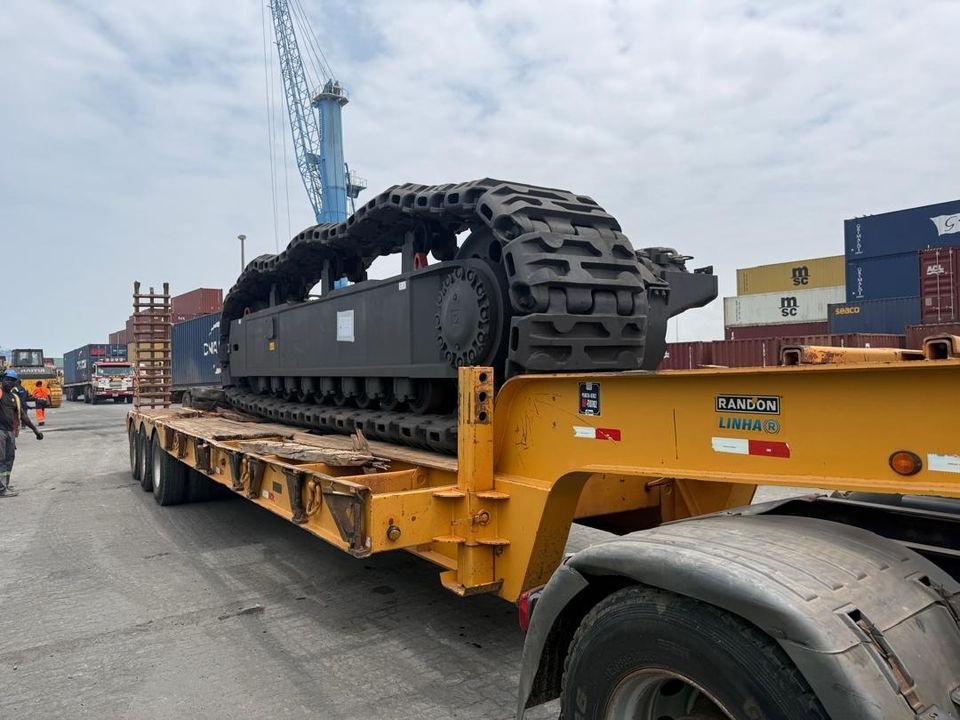 Crawler track frame on a yellow lowbed trailer in a container terminal, with a harbour crane behind