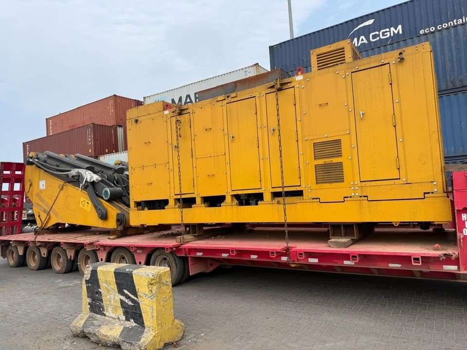 Yellow enclosed equipment module and a boom section chained down on a multi-axle lowbed trailer beside stacked containers