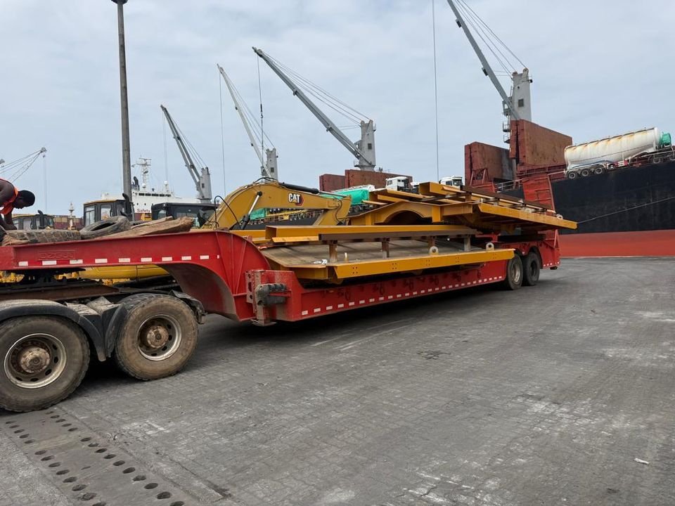 Yellow steel sections loaded on a red lowbed trailer at the quayside beside a cargo vessel, with ship cranes overhead